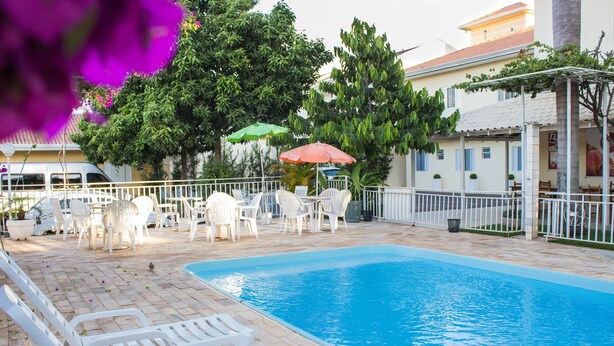 Una piscina con lettini bianchi e un tavolo con sedie, circondata da un recinto bianco e alberi.