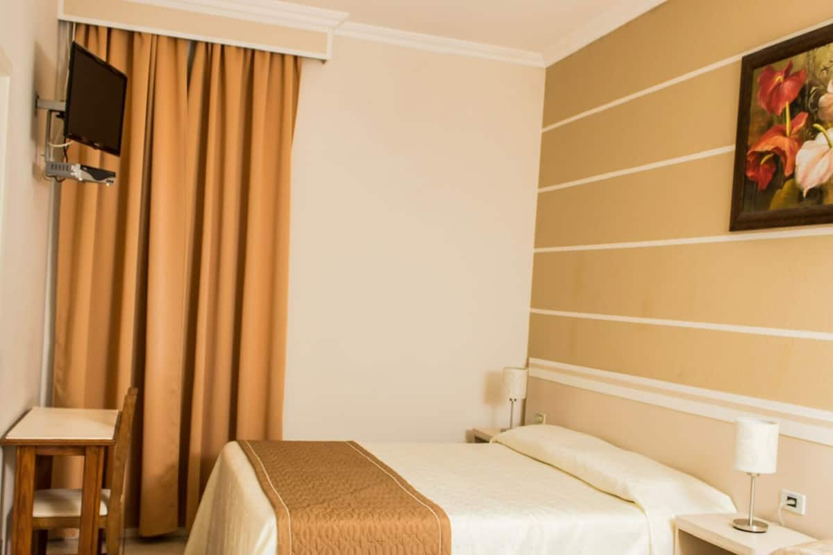 A hotel room with a bed, bedside table, lamp, TV, and a painting on the wall.