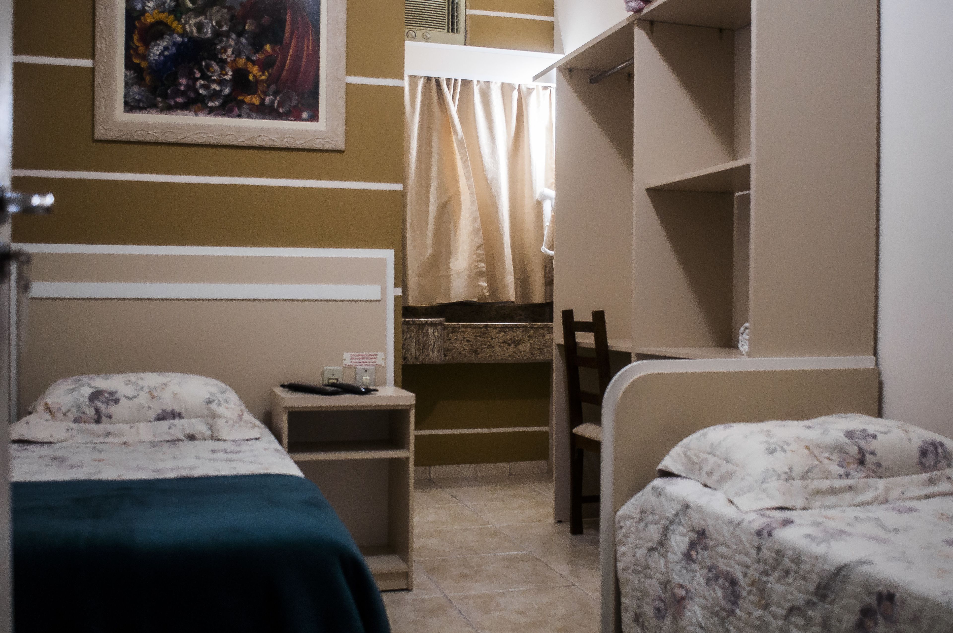 A room with two beds, a desk, a chair, a shower area, and a painting on the wall.