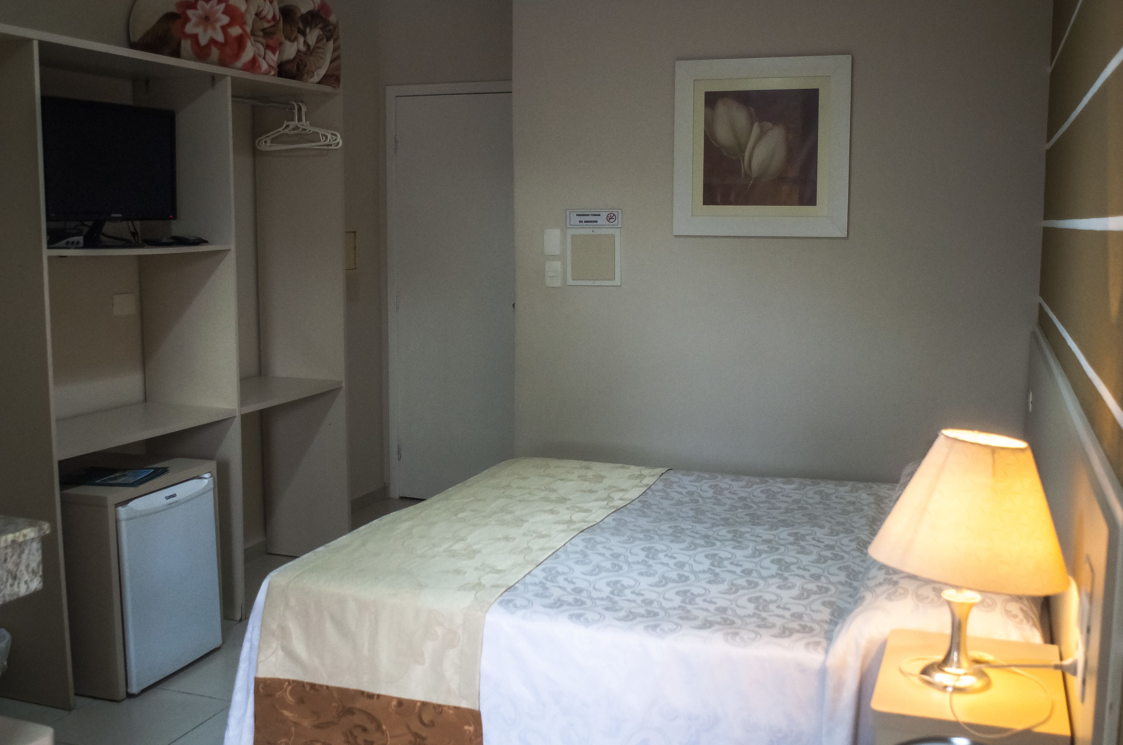 A hotel room with a bed, a bedside lamp, a television on a shelf, and a small refrigerator.