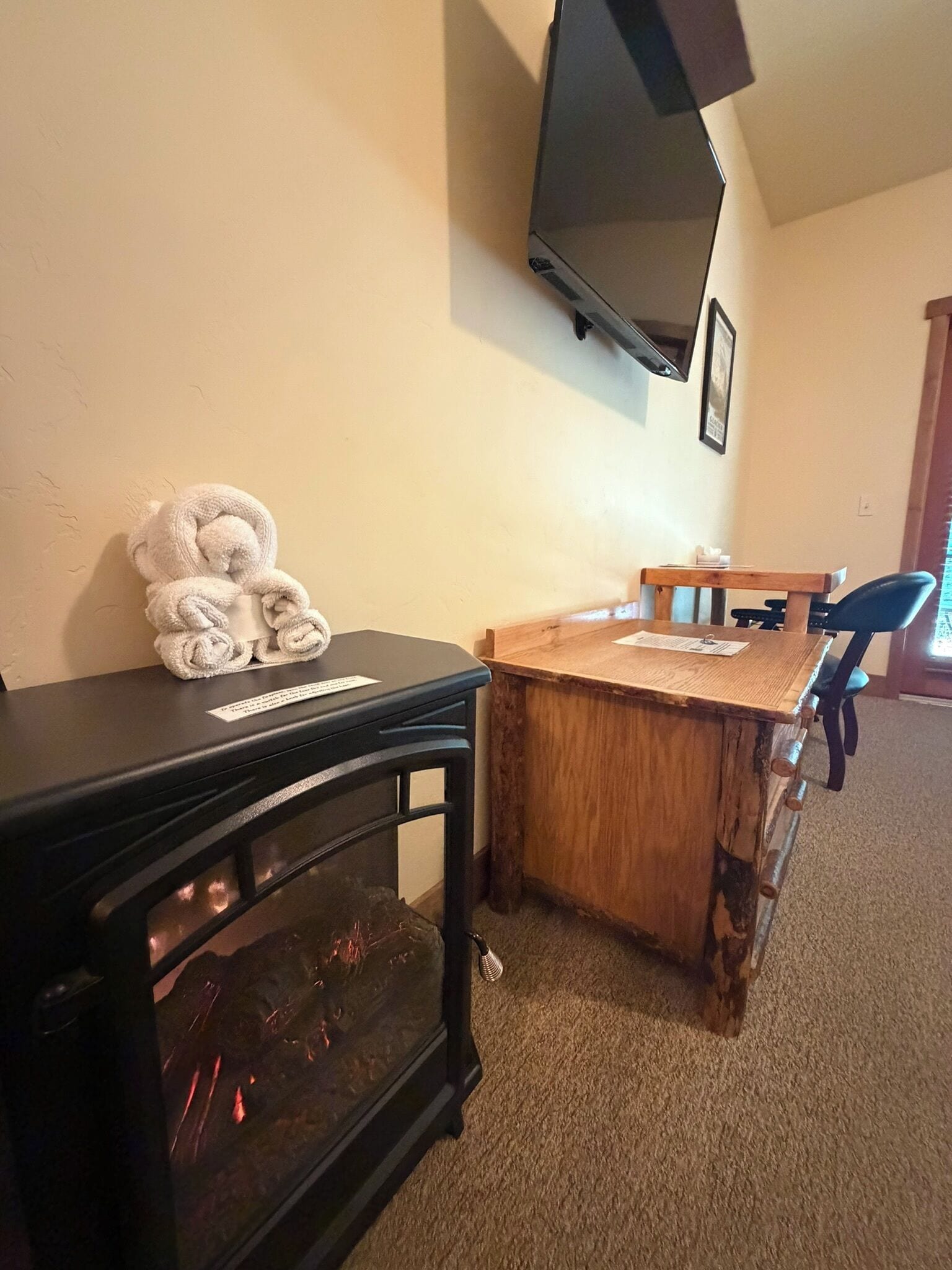 A room with a wooden dining table, a black TV mounted on the wall, and a small black table with a towel on it.