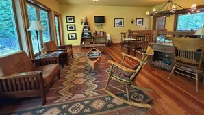 A cozy living room with a fireplace, wooden furniture, and a large window overlooking trees. - Glacier Guides Lodge (West Glacier)