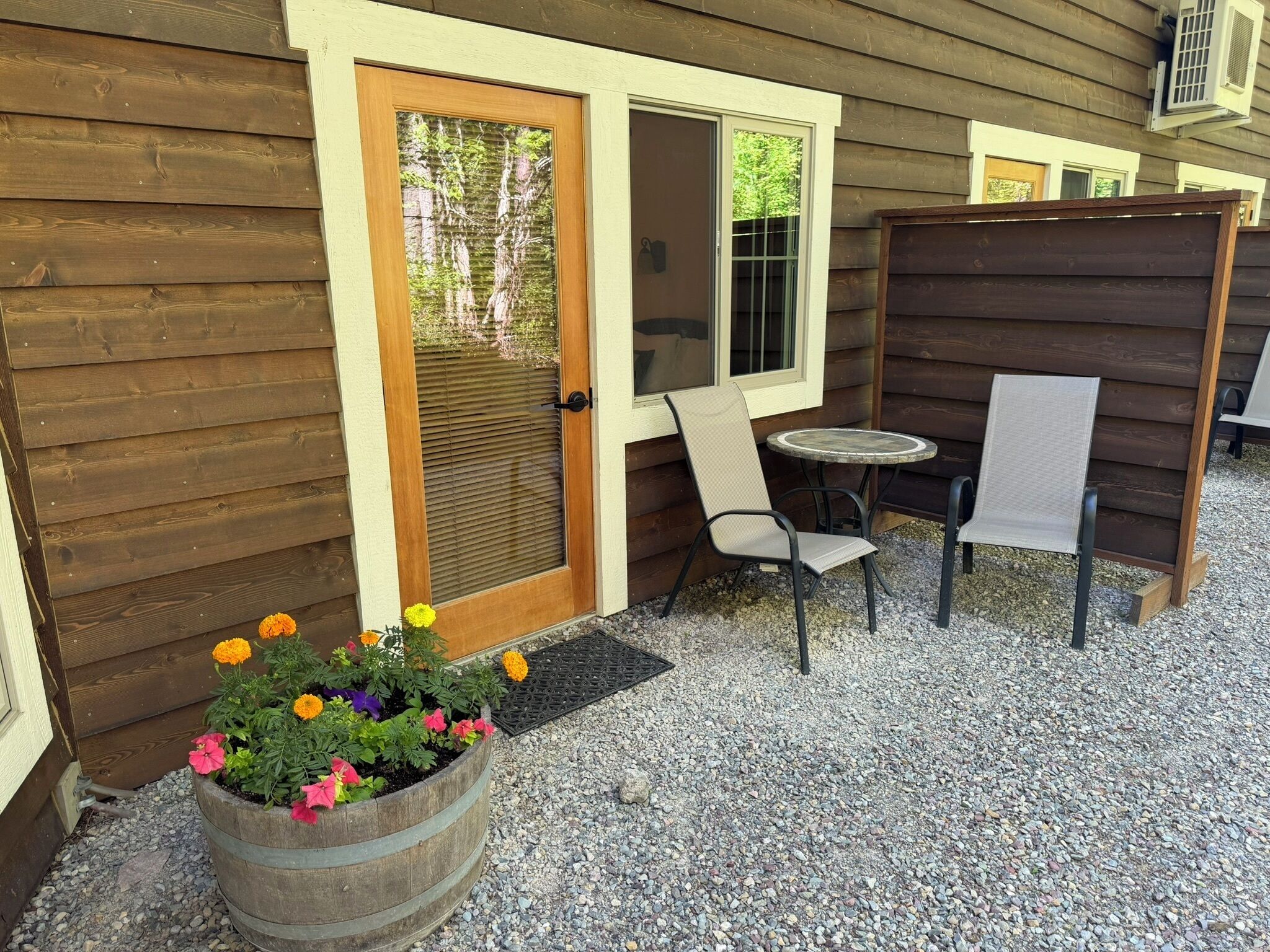 A wooden house with a door, a window, a small patio with a table and chairs, and a flower pot with flowers.