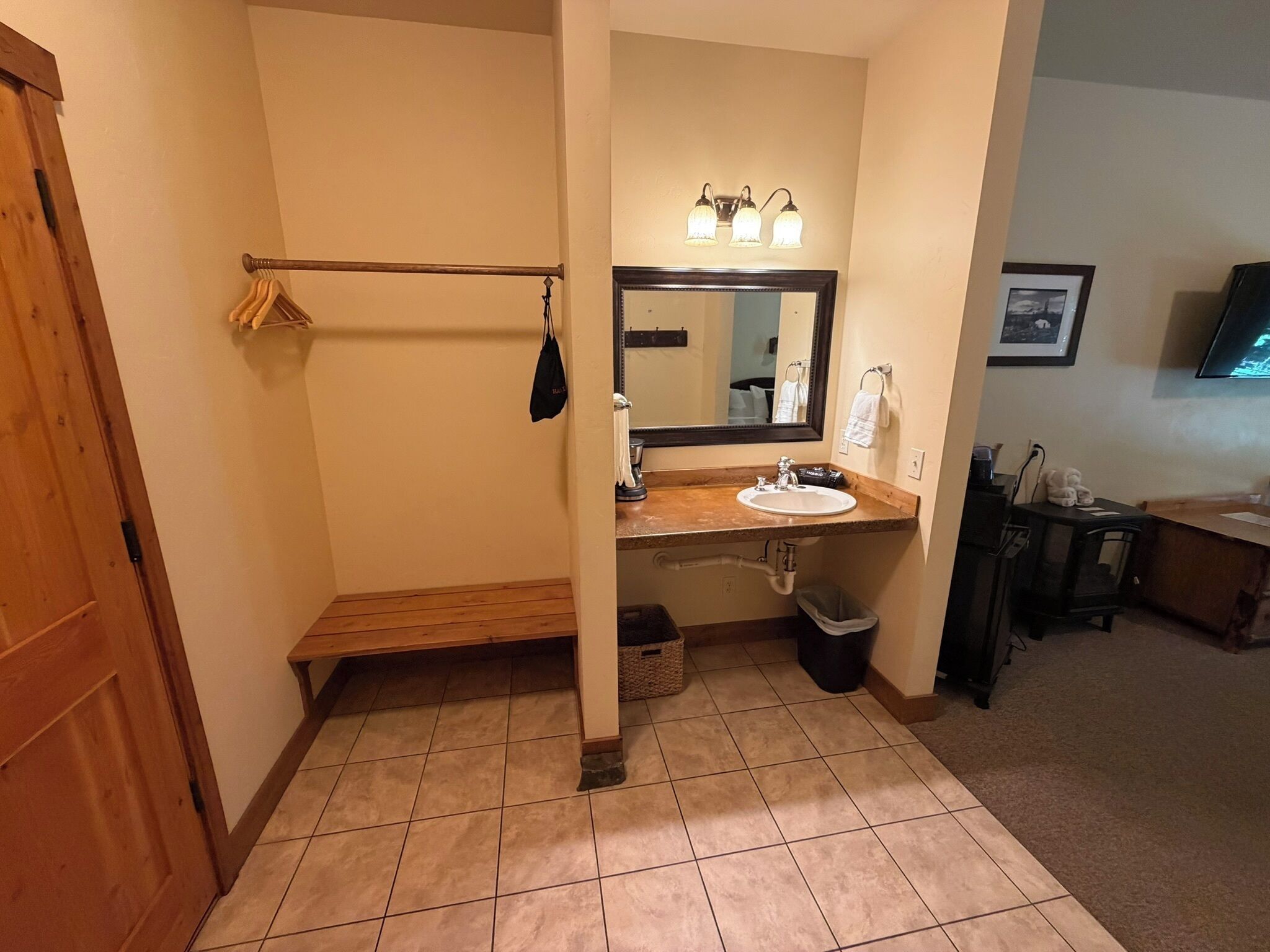 A bathroom with a wooden bench, a sink, a mirror with lights, and a trash bin.