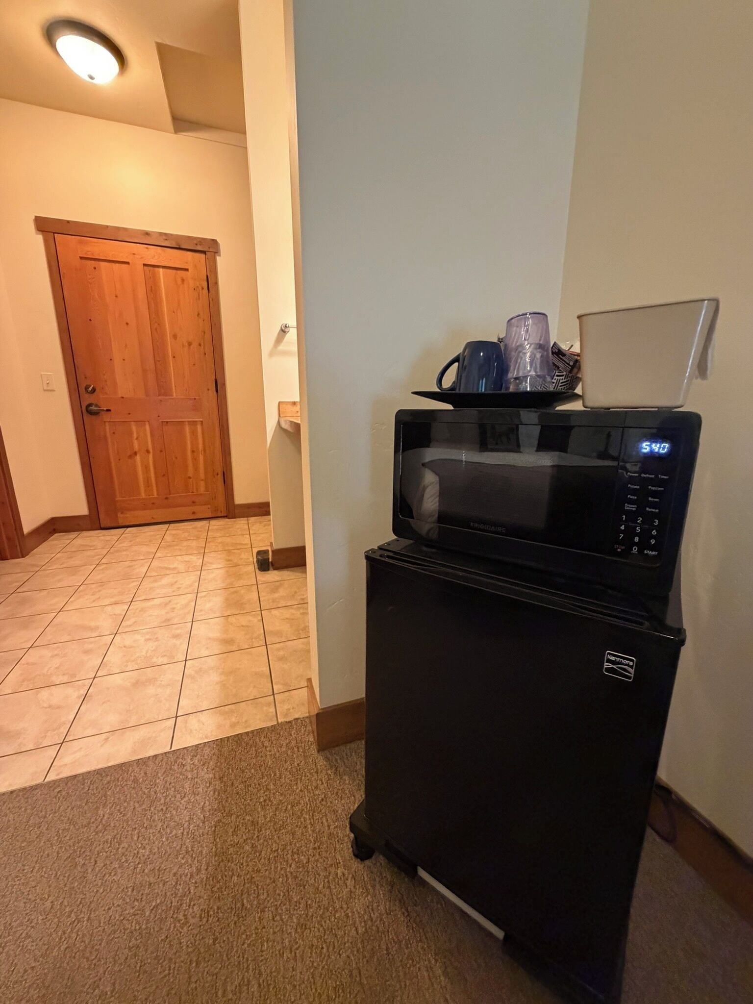 Una cocina con un microondas sobre carrito, una puerta de madera y suelo de baldosas.