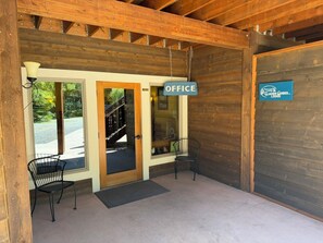 A wooden lodge entrance with a sign labeled 'OFFICE,' two chairs outside, and a glass door leading to a staircase.