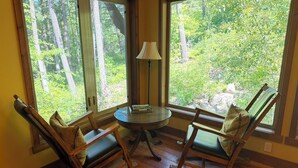 A room with two rocking chairs, a small table, and a lamp. Large windows offer a view of a forest. - Glacier Guides Lodge (West Glacier)