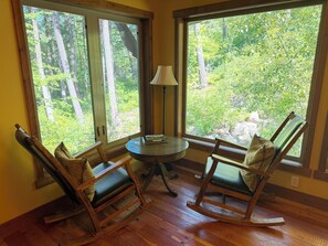 A room with two rocking chairs, a small table, and a lamp. Large windows offer a view of a forest.