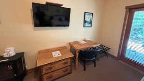 A room with a flat-screen TV mounted on the wall, a small wooden table with a chair, a heater, and a window with blinds. - Glacier Guides Lodge (West Glacier)