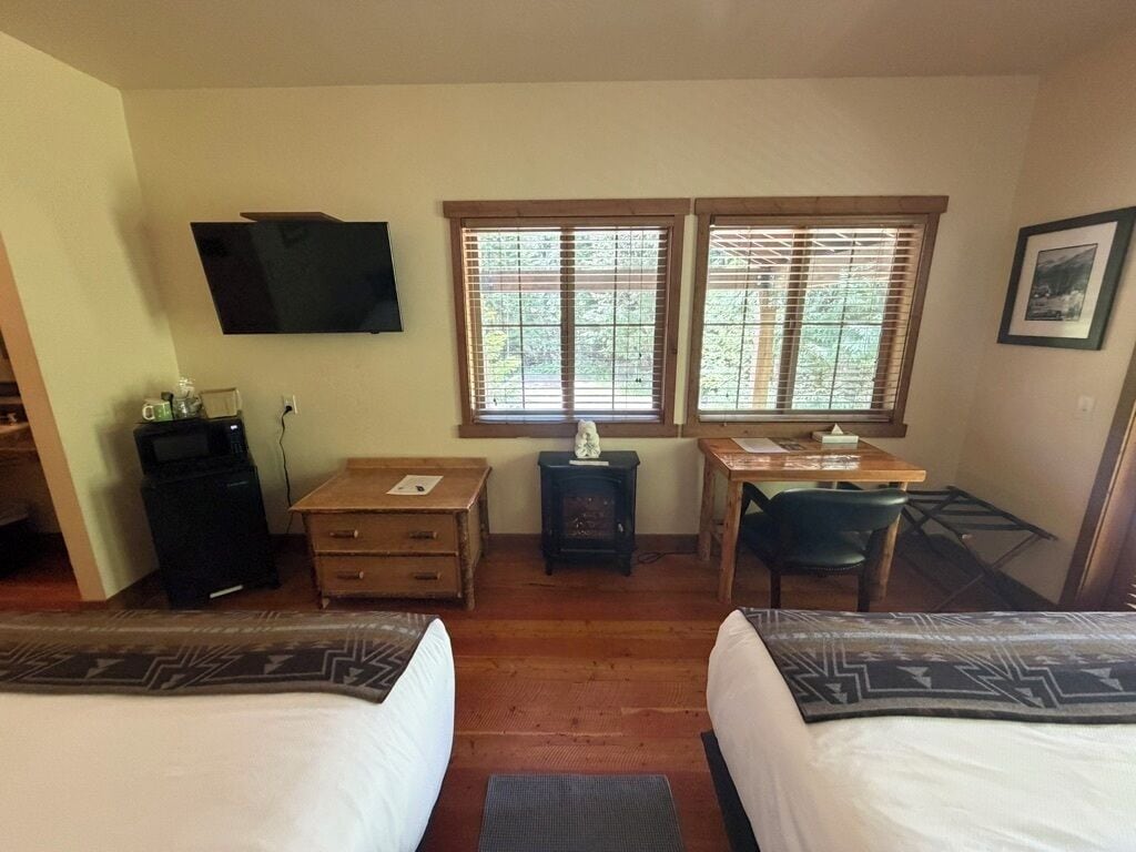 A hotel room with two beds, a desk, a chair, a television, and windows with blinds.
