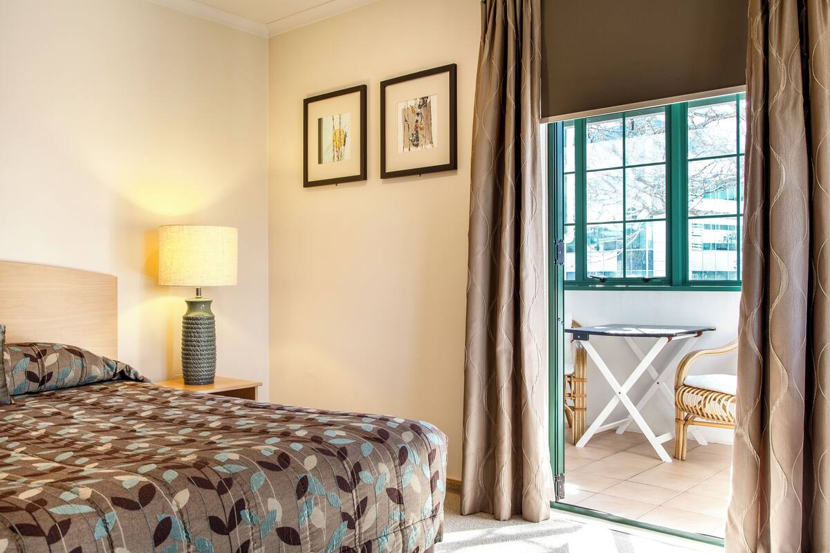 A hotel room with a bed, a lamp, two framed pictures on the wall, and a view of the outside through a window.