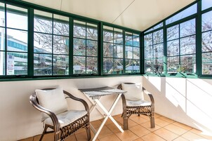A conservatory with wicker furniture, a small table, and a view of a building and trees through large windows.