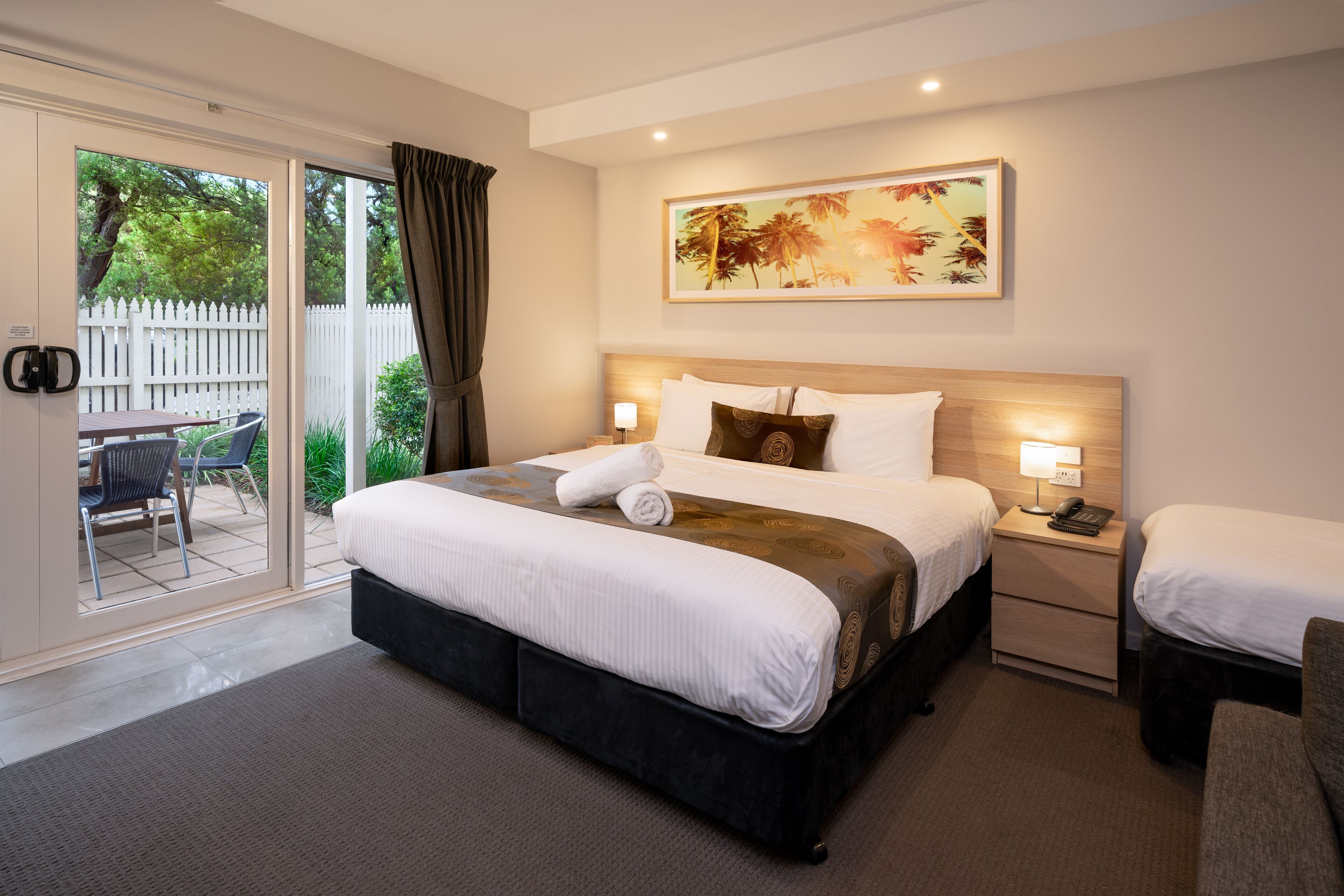 Habitación de hotel con una cama grande, una mesita de noche con lámpara, una silla y un ventanal corredero que da a un espacio exterior con mesa y sillas.