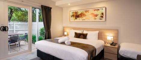 Habitación de hotel con una cama grande, una mesita de noche con lámpara, una silla y un ventanal corredero que da a un espacio exterior con mesa y sillas.
