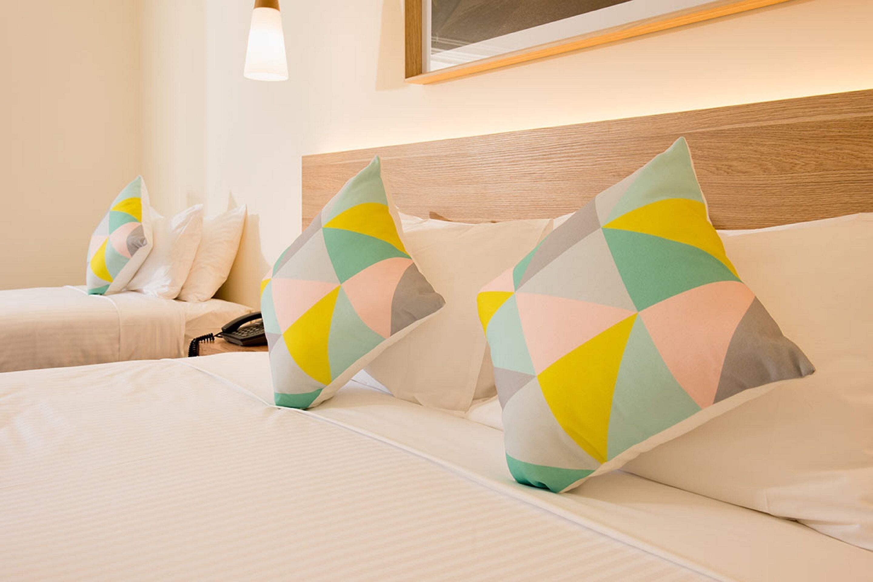Une chambre d’hôtel avec une tête de lit en bois, un lit recouvert de draps blancs et des coussins aux motifs géométriques et aux couleurs vives.