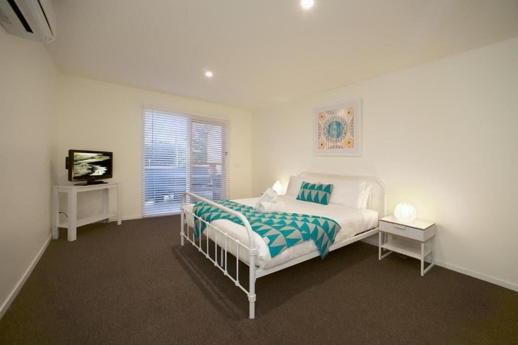 A bedroom with a white metal bed frame, a teal and white bedspread, a bedside table with a lamp, and a window with blinds.