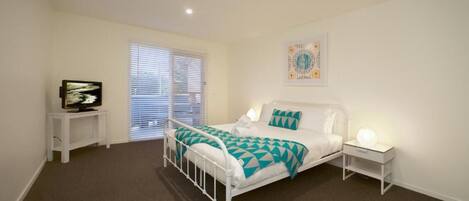 Un dormitorio con estructura de cama de metal blanco, colcha blanca y azul aguamarina, mesita de noche con lámpara y ventana con persianas.