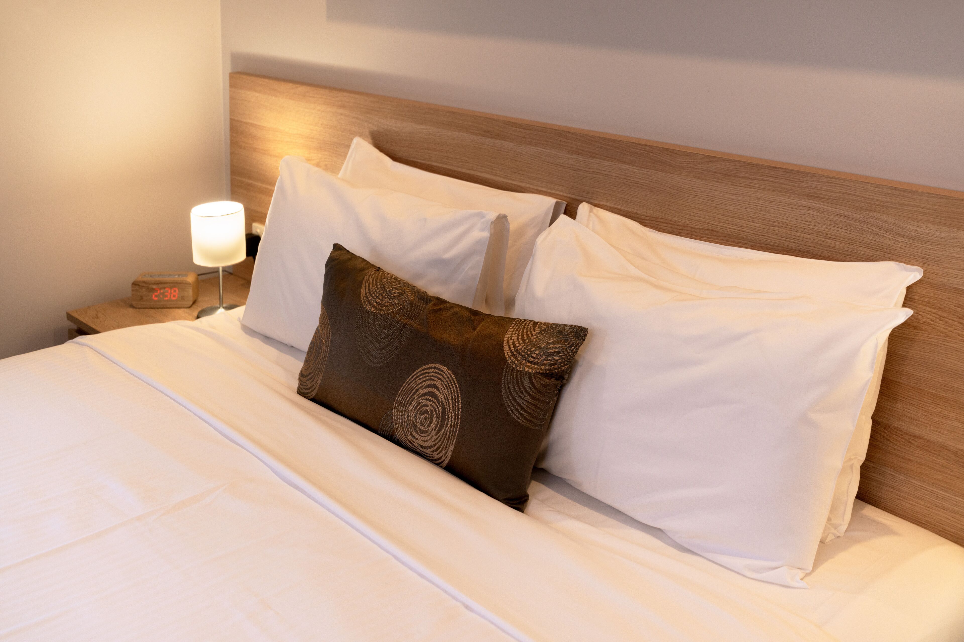 A neatly made bed with white linens and a brown patterned pillow, a wooden headboard, and a bedside table with a lamp and a clock.