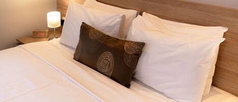 A neatly made bed with white linens and a brown patterned pillow, a wooden headboard, and a bedside table with a lamp and a clock.