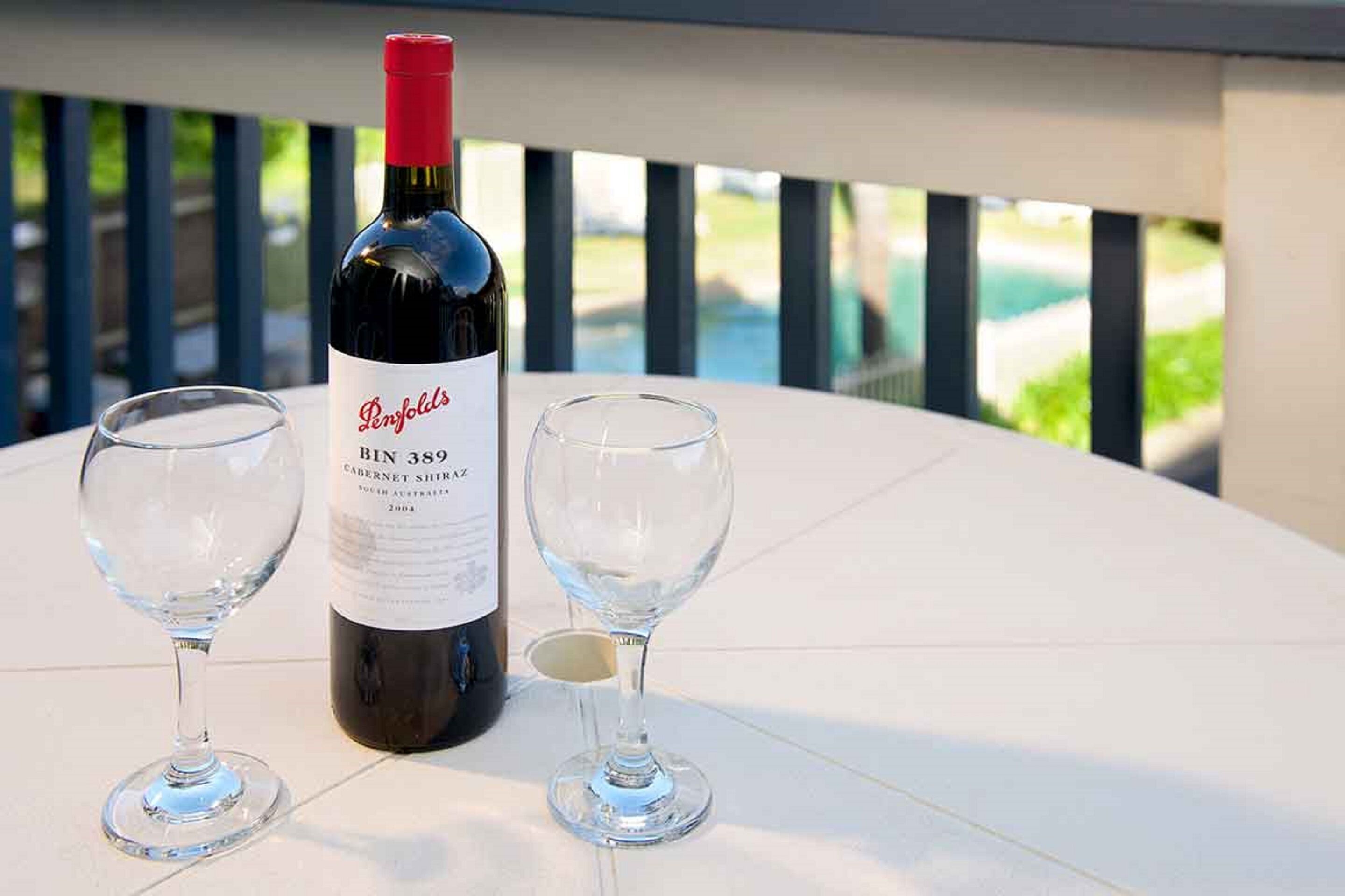 Eine Flasche Penfolds Bin 3896 Cabernet Sauvignon und zwei Weingläser auf einem weißen Tisch, im Hintergrund Grün und ein Pool zu sehen.
