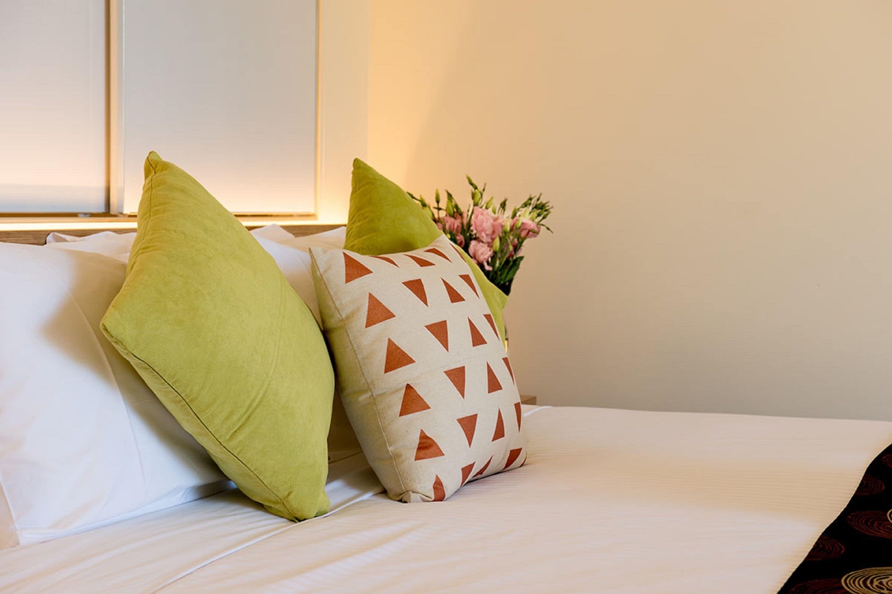 A bed with two pillows, one green and one with a geometric pattern, and a vase with flowers in the background.