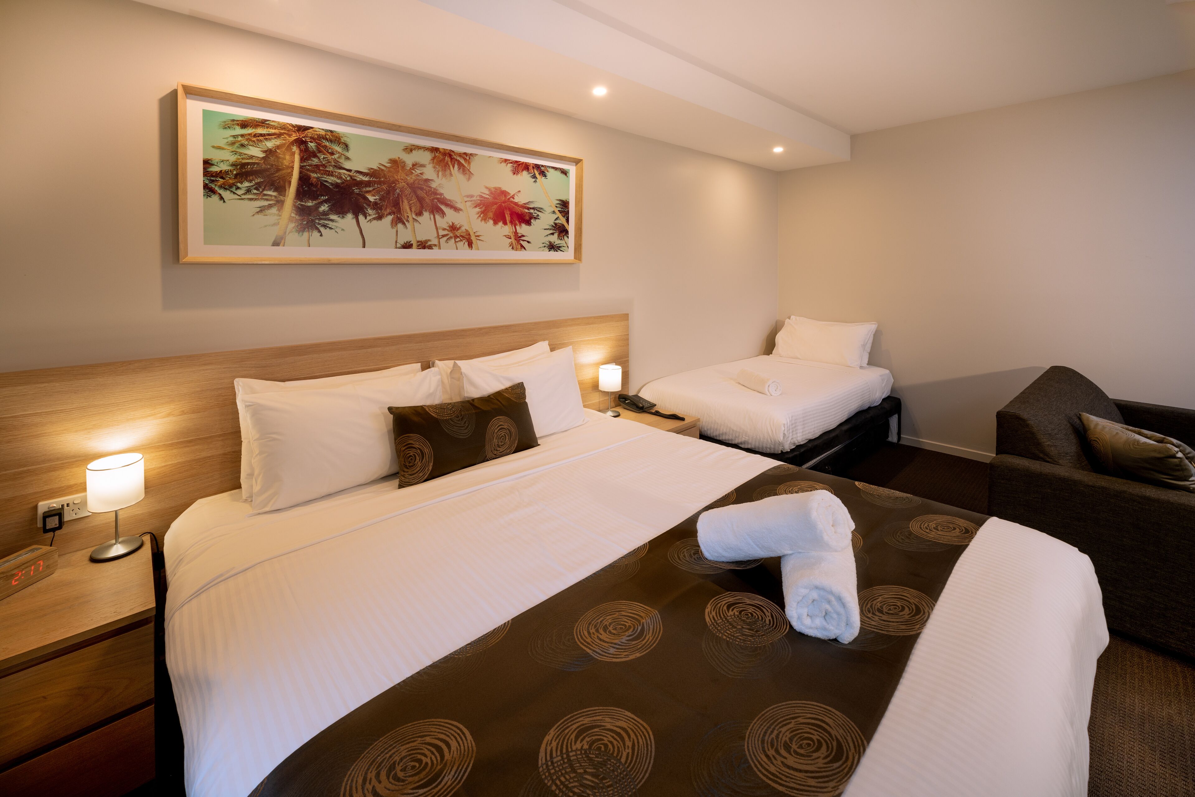 A hotel room with a large bed, two bedside tables, a nightstand, a sofa, and a painting of palm trees.