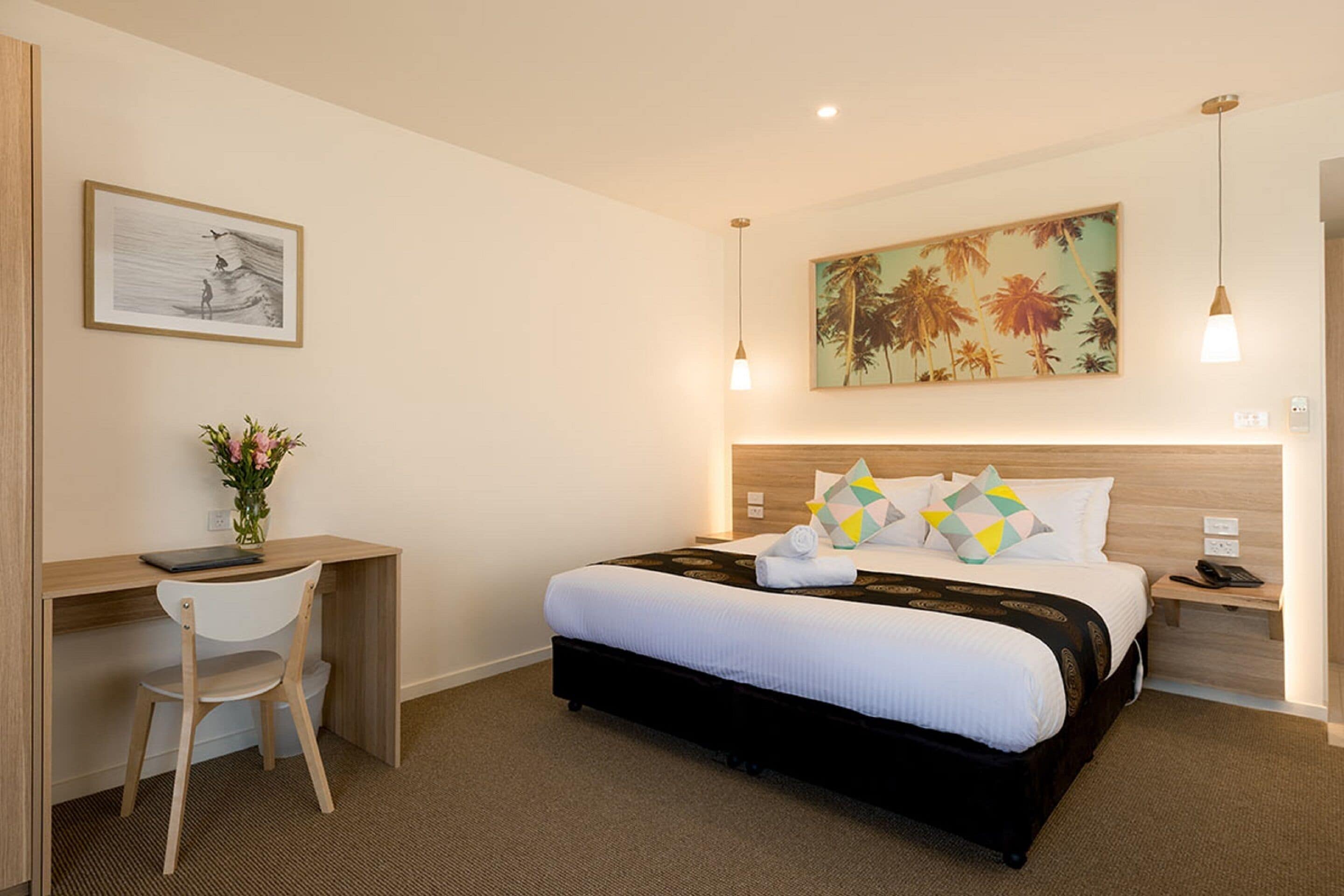 Ein Hotelzimmer mit einem Bett, einem Schreibtisch und einem Sessel. An der Wand hängt ein Gemälde und auf dem Schreibtisch steht eine Vase mit Blumen.