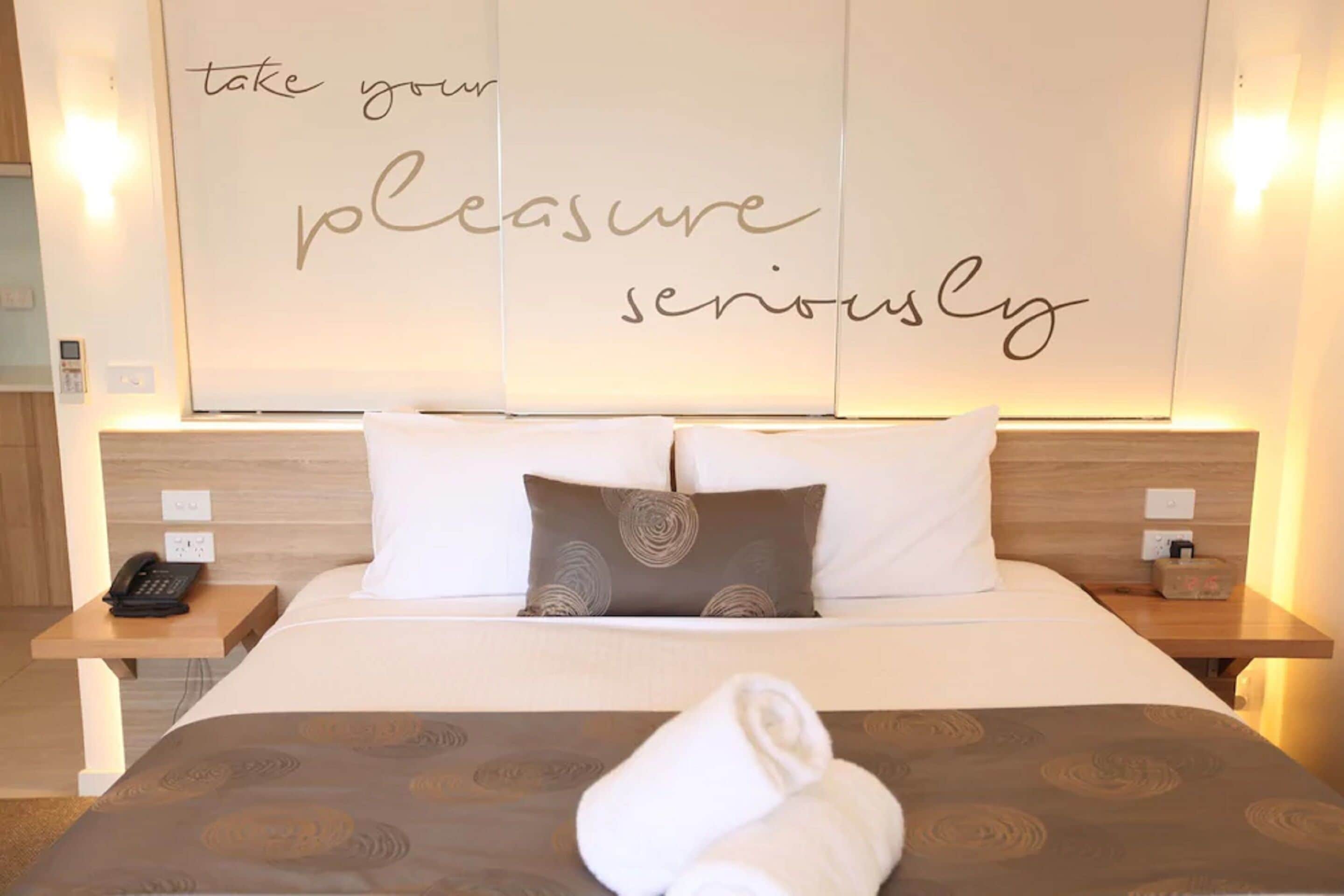A hotel room with a bed, two pillows, a bedside table with a phone, and a wall with motivational text.