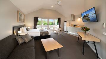 Habitación de hotel moderna con cama, sofá, mesa de comedor, televisor y balcón con vistas a zonas verdes.