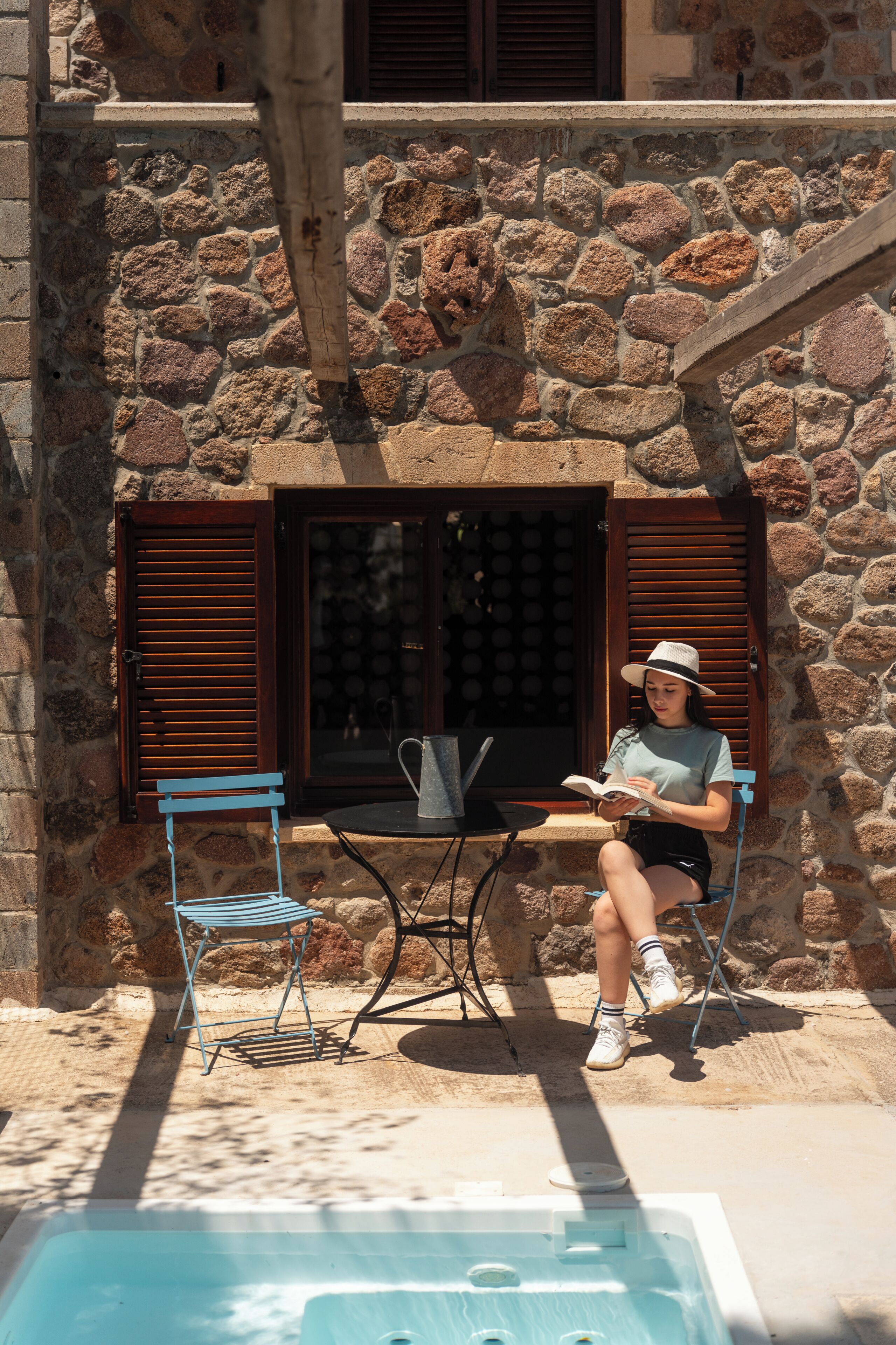 Un bâtiment en pierre avec une piscine, une personne assise sur une chaise en train de lire un livre, et une table avec une chaise.