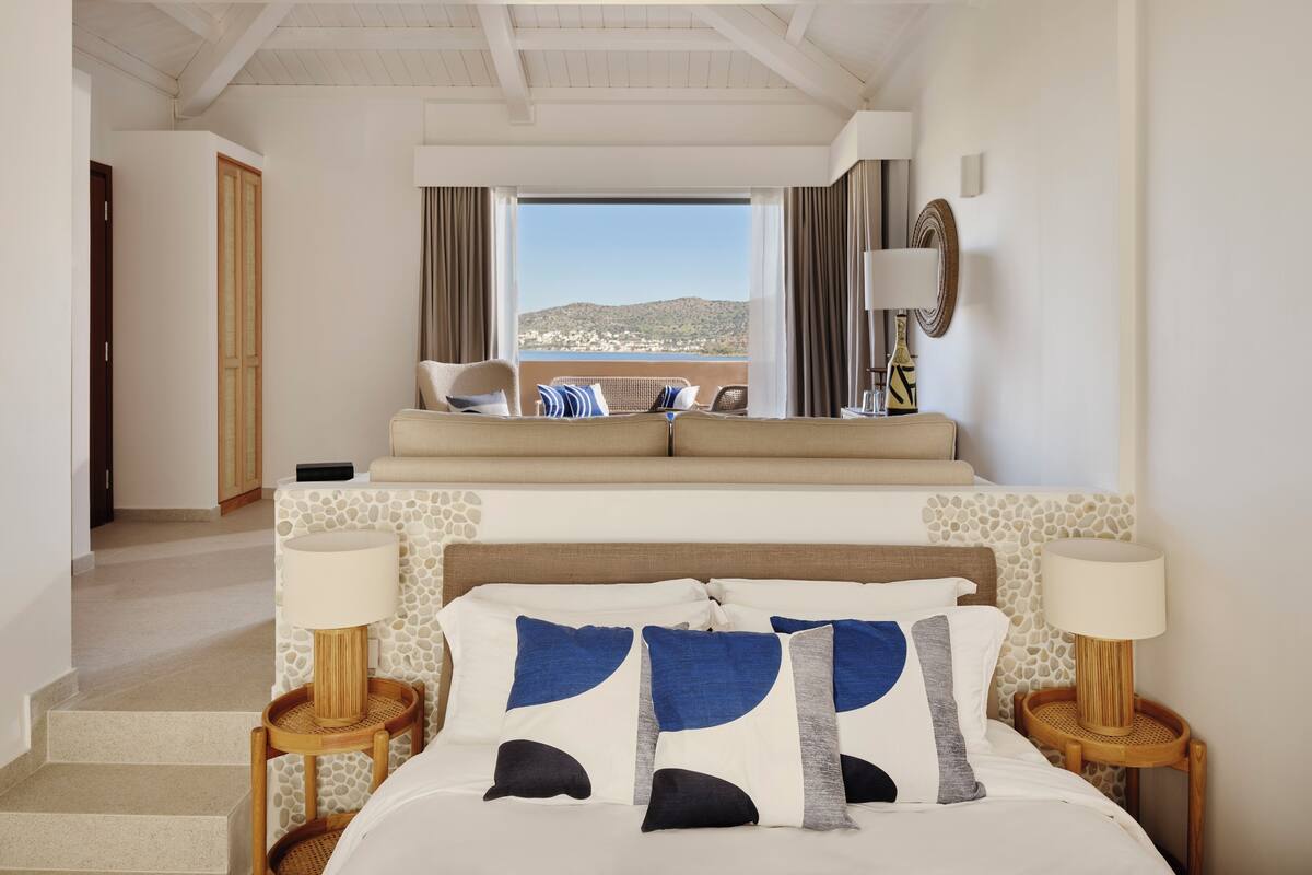 A hotel room with a bed, two bedside tables, a sofa, a view of the sea, and a staircase leading to another room.
