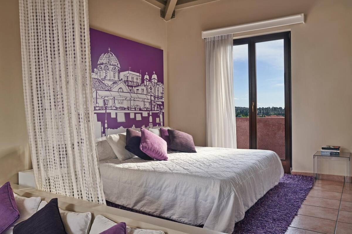 A bedroom with a bed, a purple wall mural, a large window, and a view of the landscape.