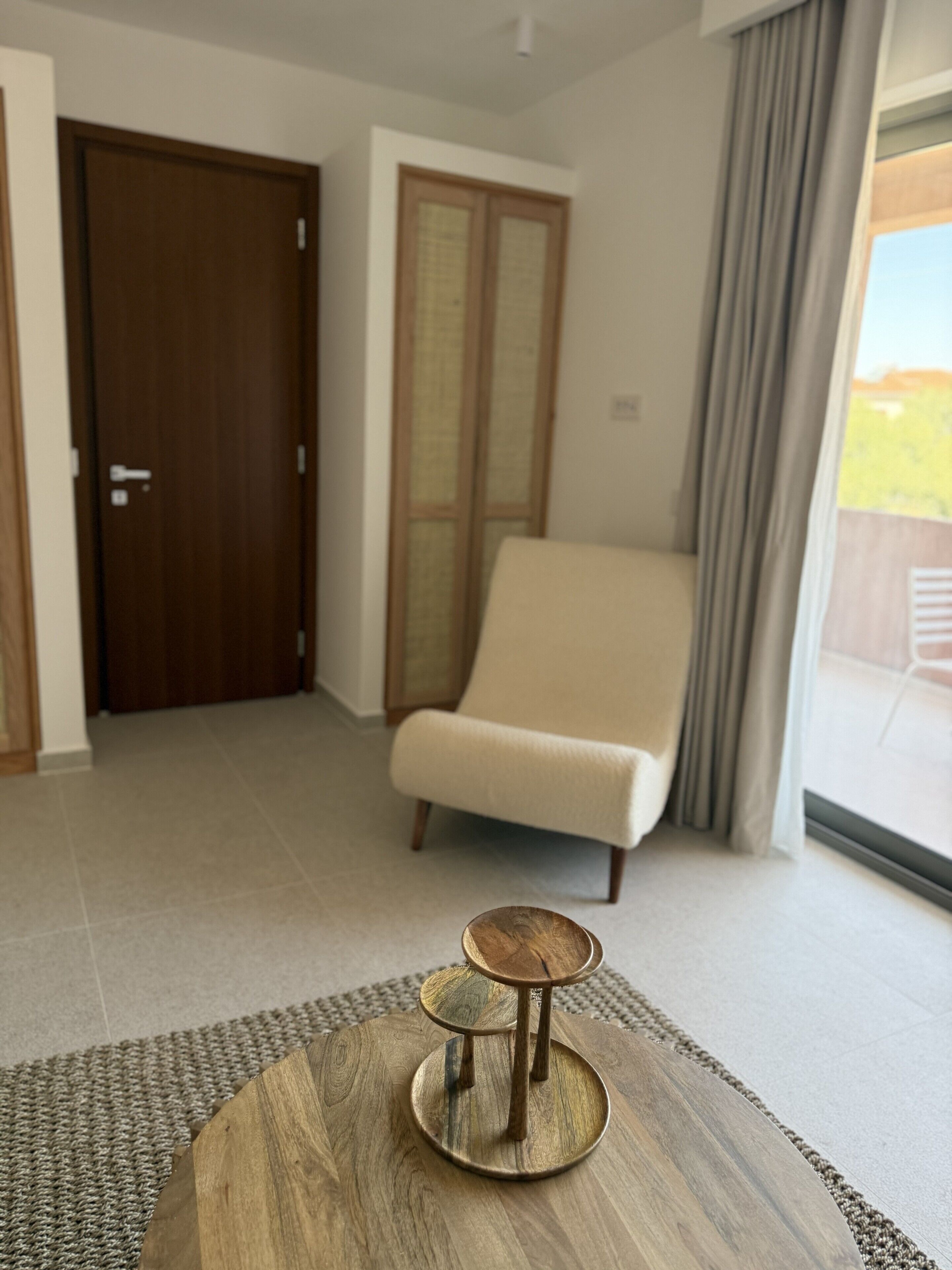 A modern living room with a wooden coffee table, a beige armchair, and a large window with curtains.