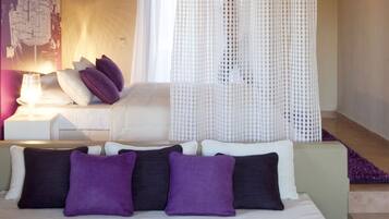 A hotel room with a bed, a white sofa, a small table with a vase and fruit, and a clear glass table.