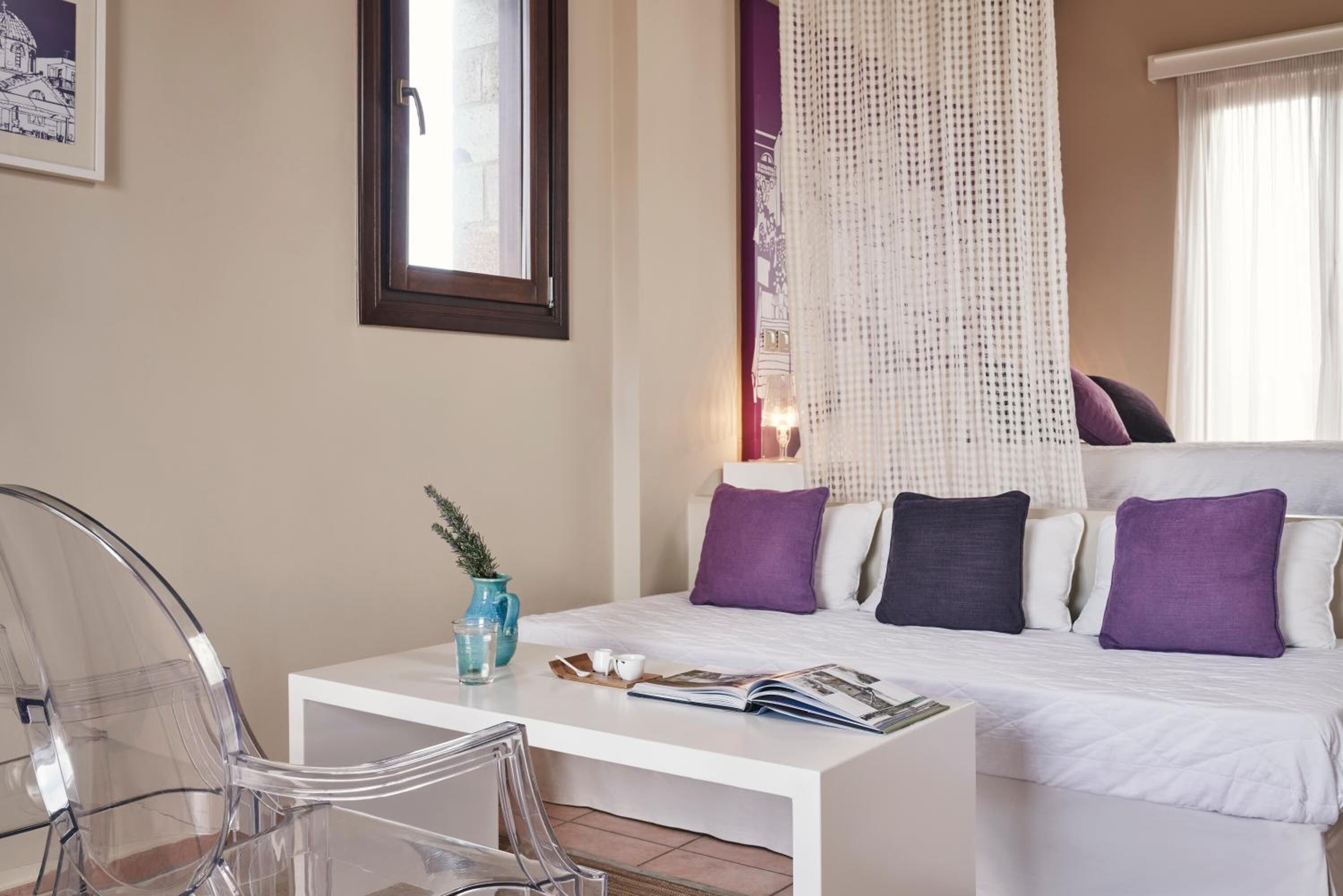 Une chambre d’hôtel avec un lit, une table de chevet blanche, une chaise transparente et une fenêtre donnant sur un immeuble.