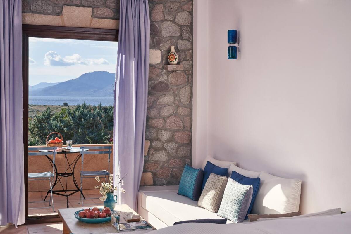 A room with a stone wall, a balcony with a table and chairs, and a bed with pillows.