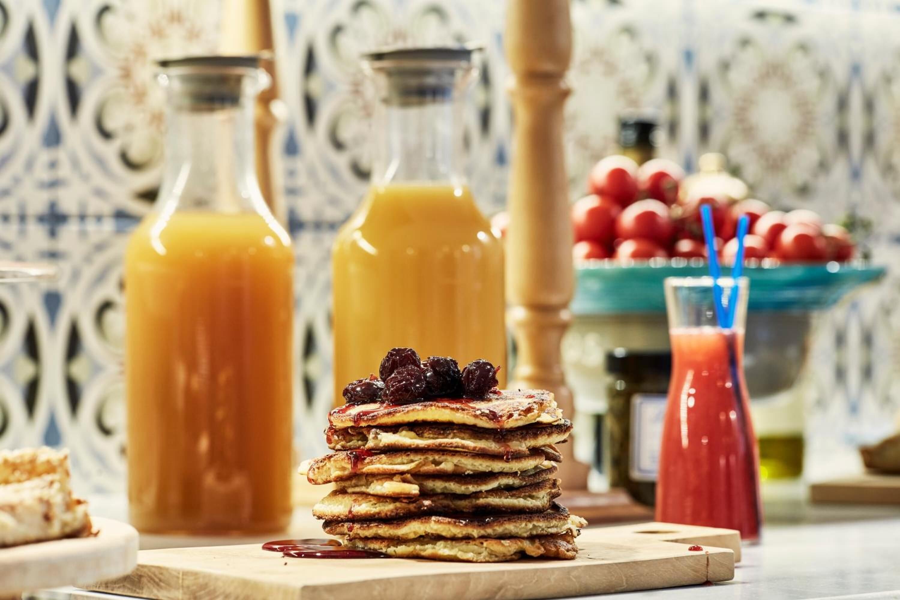 A stack of pancakes with syrup and berries, a glass of orange juice, and a glass of red juice on a table.