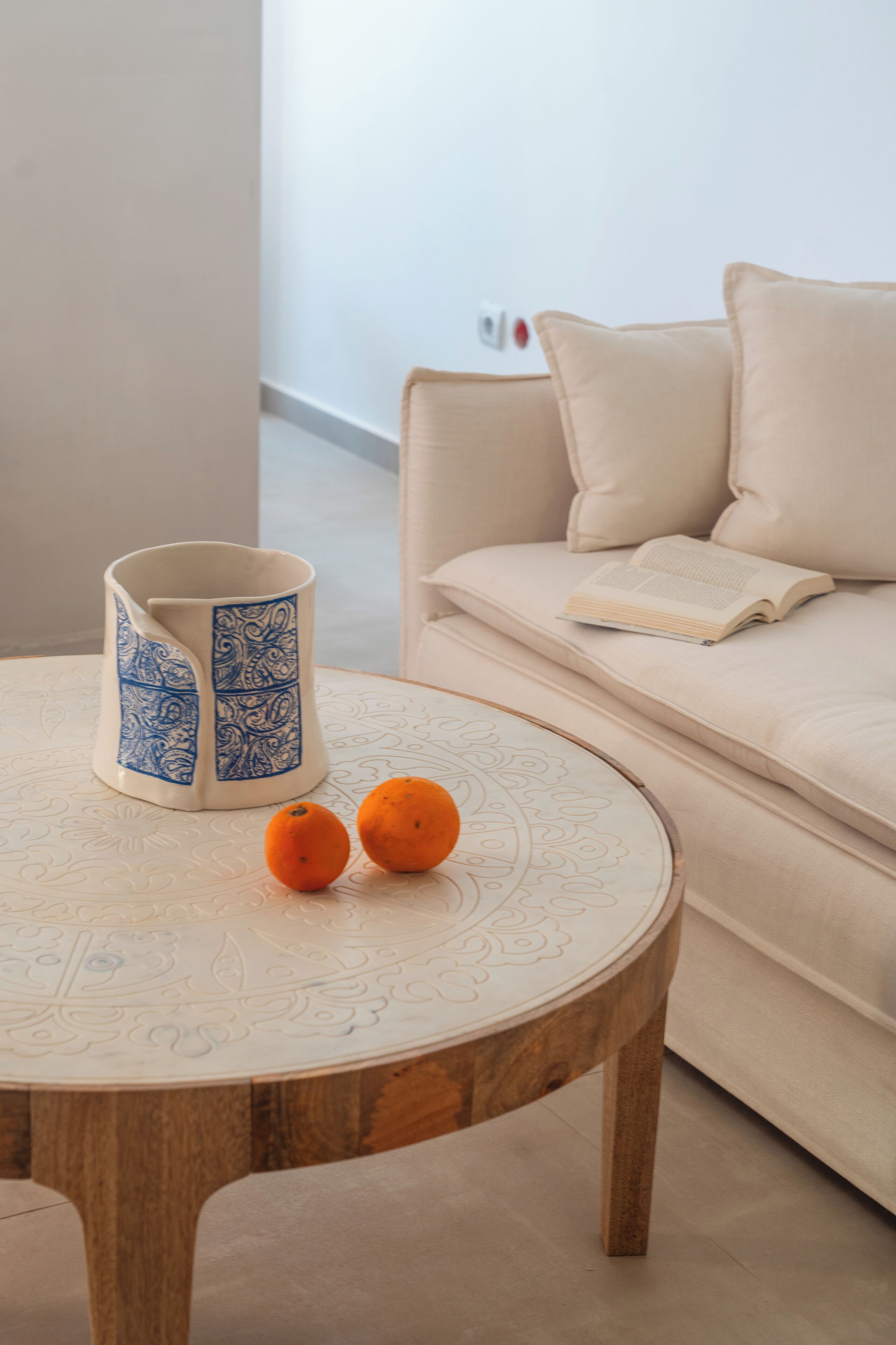 A round wooden table with a decorative white top, a blue and white patterned mug, and two oranges.