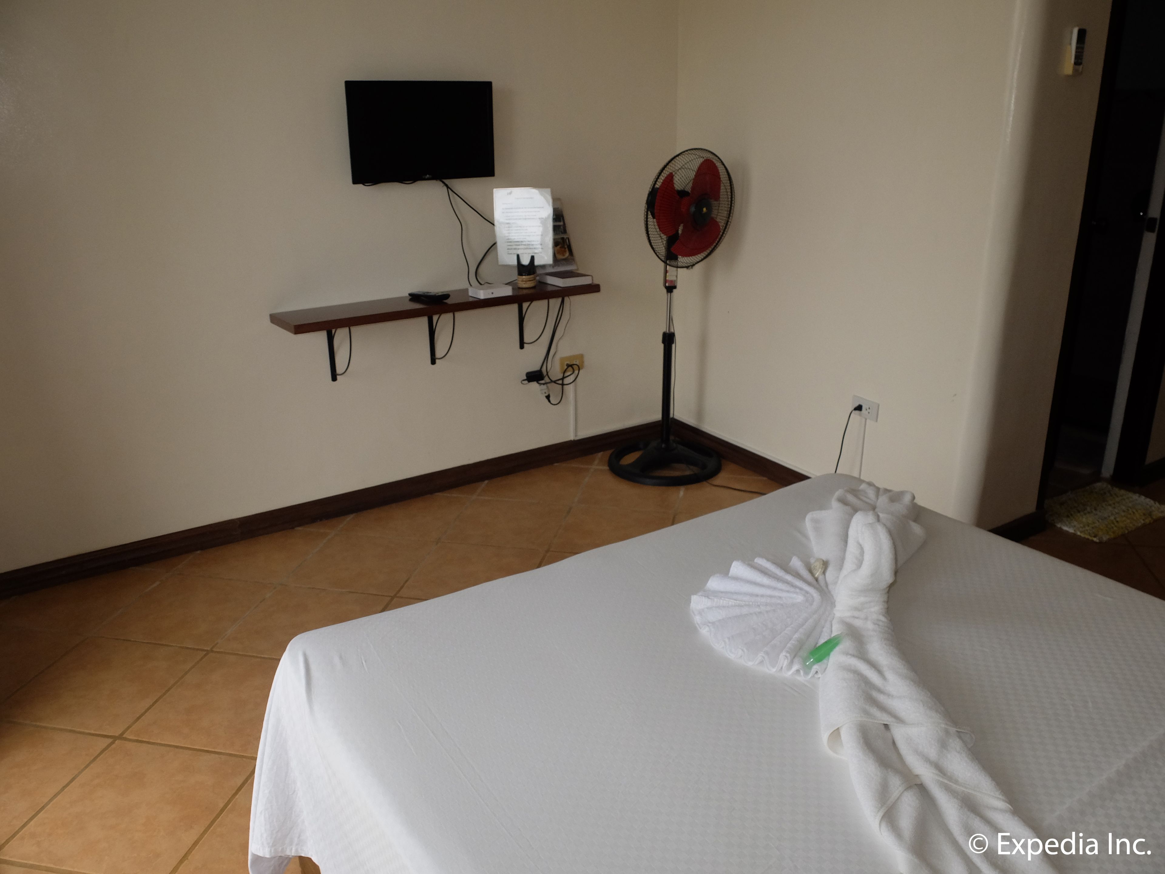 A hotel room with a bed, a flat-screen TV mounted on the wall, a desk with a computer and a fan, and a tiled floor.