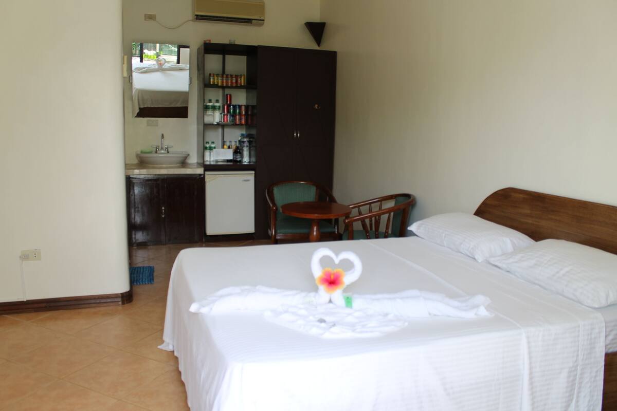 A hotel room with a bed, a small desk, a chair, a kitchenette, and a wardrobe.