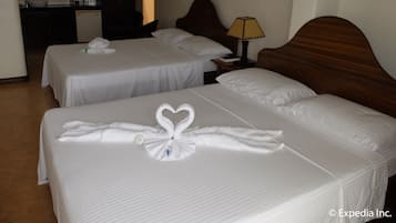 A hotel room with two beds, each with a heart-shaped towel arrangement. There is a wooden headboard, a bedside lamp, and a painting on the wall.