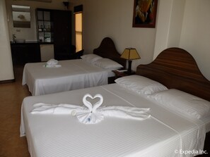 A hotel room with two beds, each with a heart-shaped towel arrangement. There is a wooden headboard, a bedside lamp, and a painting on the wall.