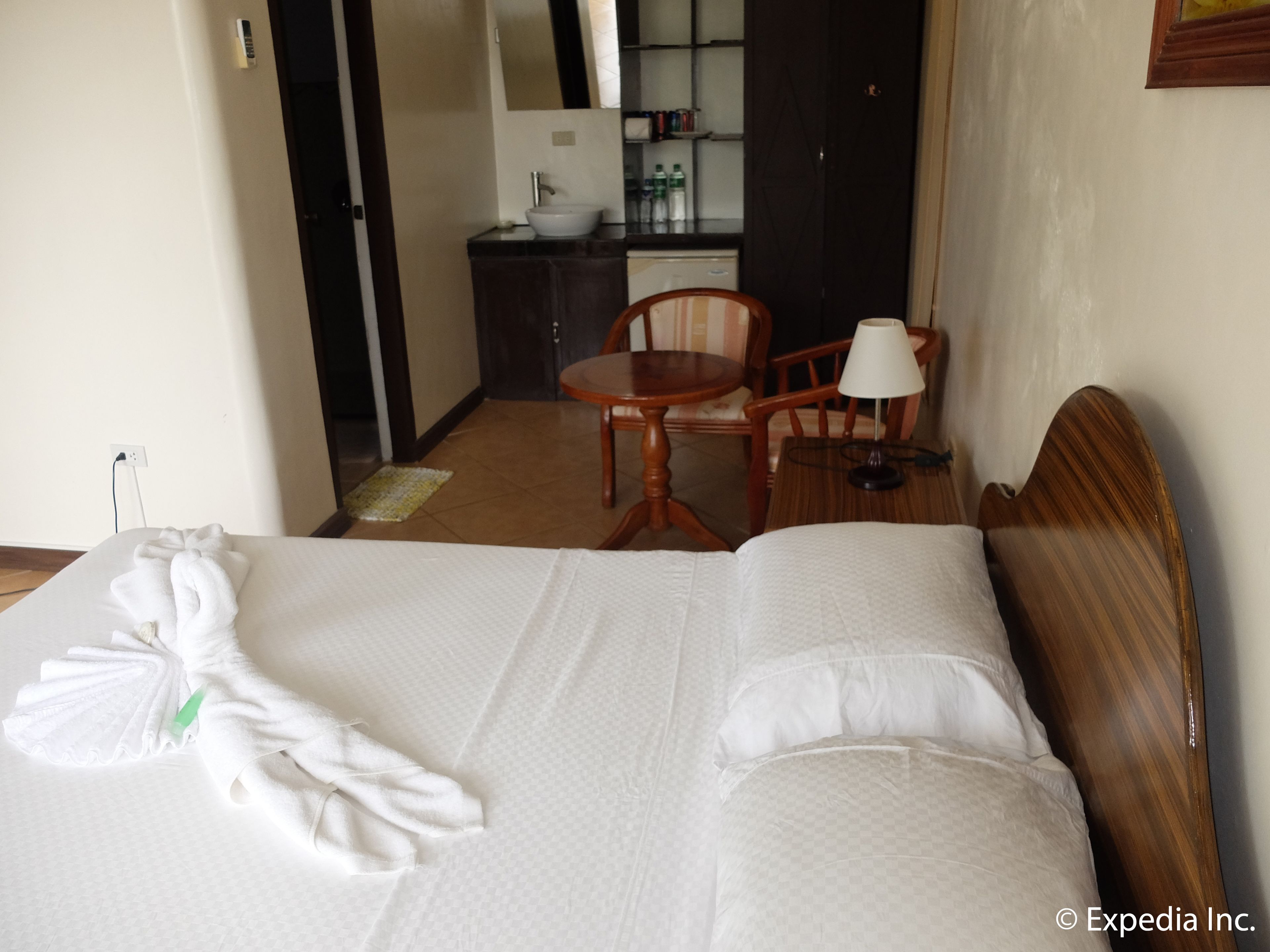 A hotel room with a neatly made bed, a wooden chair, a small table with a lamp, and a bathroom visible through an open door.