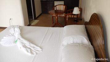 A hotel room with a neatly made bed, a wooden chair, a small table with a lamp, and a bathroom visible through an open door.