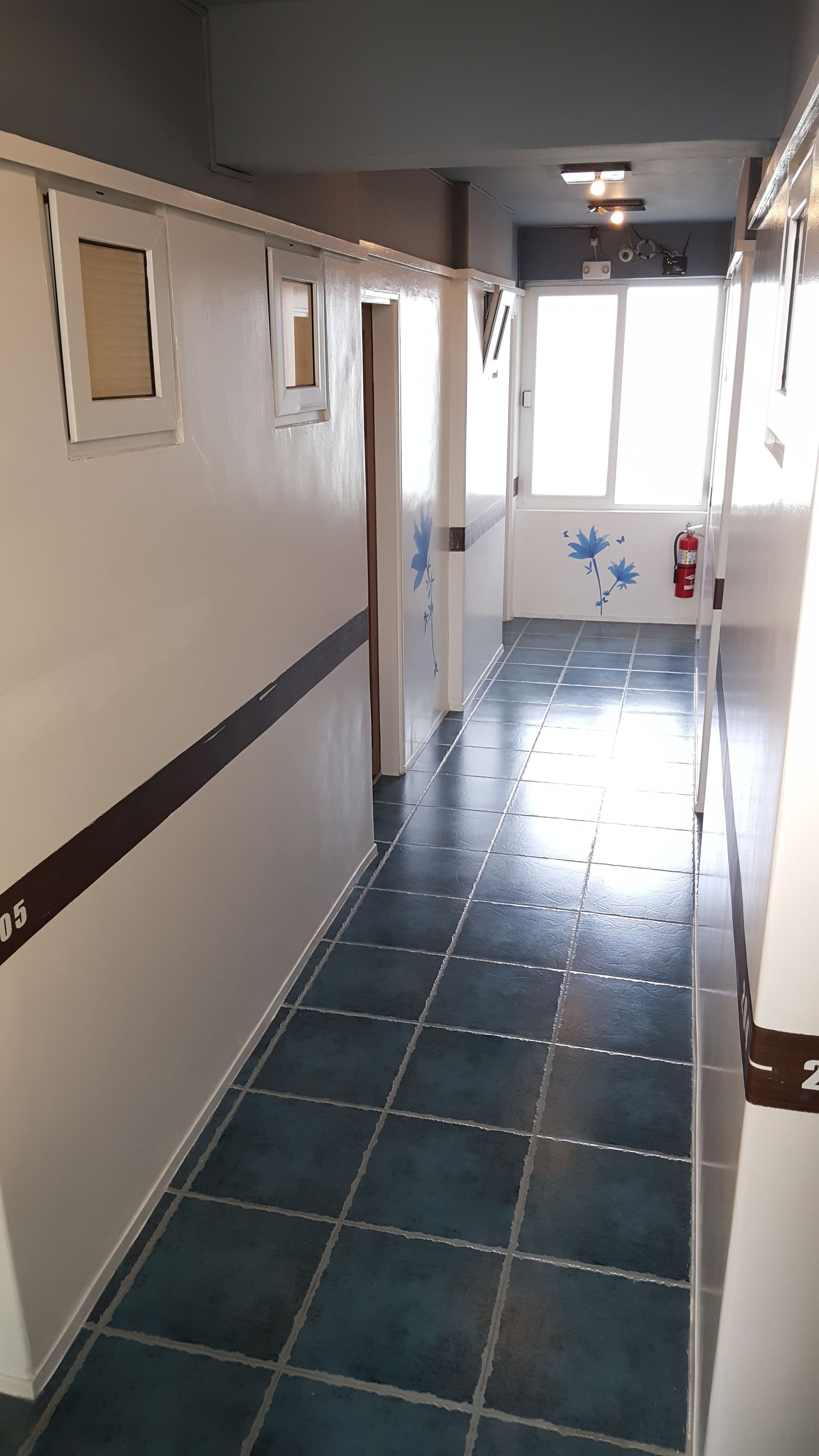 A corridor with dark blue tiled flooring, white walls with a brown stripe, and a fire extinguisher mounted on the wall.