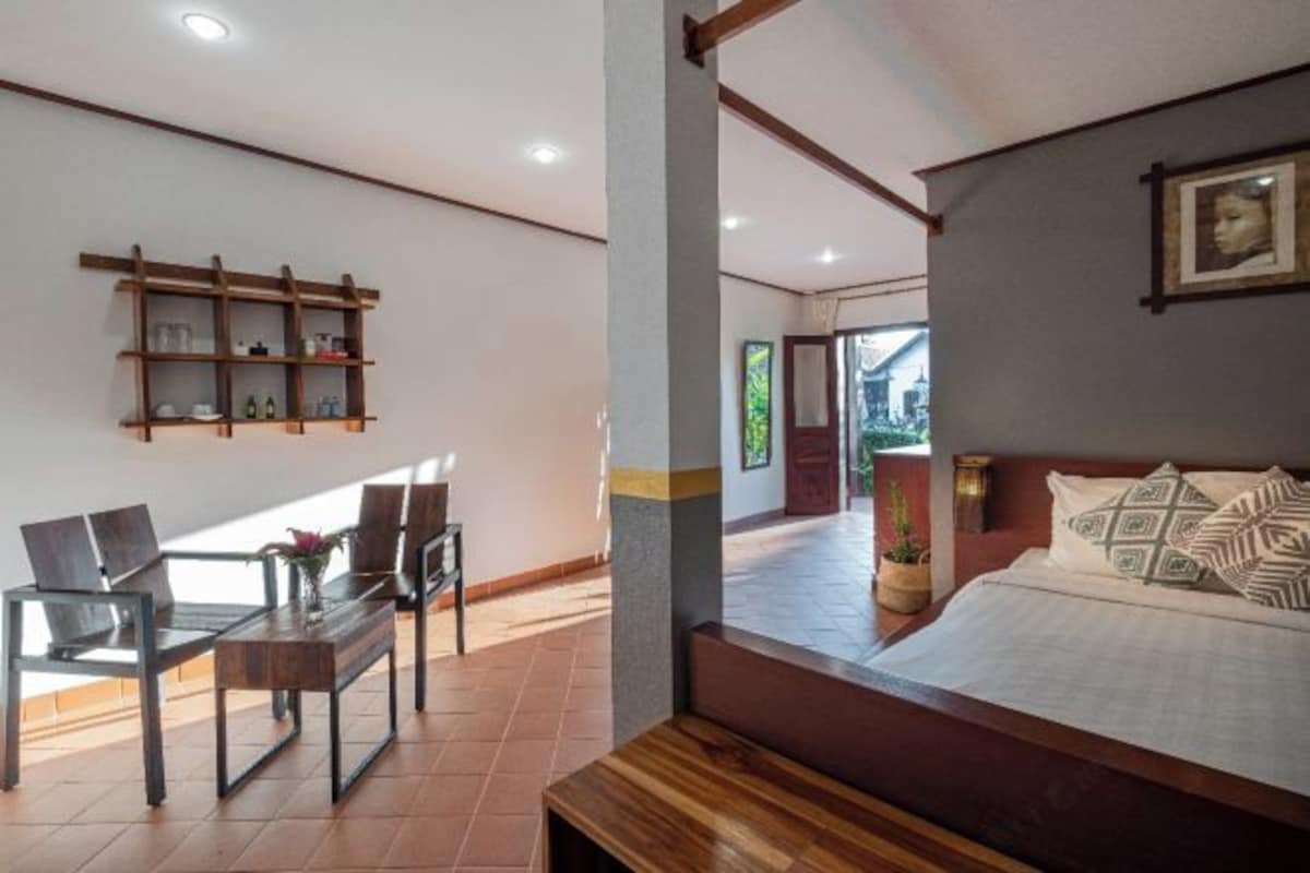 Una habitación de hotel con una cama, un banco de madera, una mesa con sillas y un estante en la pared con objetos.