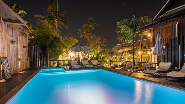 Un área de piscina nocturna con sillones y sombrillas, rodeada de edificios de madera y palmeras.