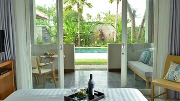 A hotel room with a bed, a dining table with chairs, a sofa, and a view of a swimming pool and garden.