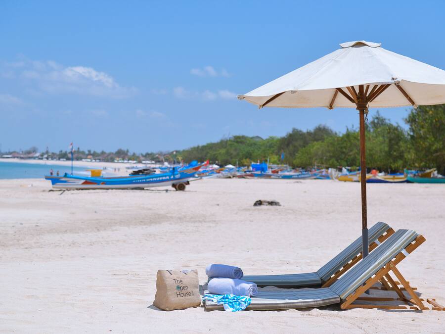 Escena de playa con una silla de descanso y una sombrilla, un bolso de playa y un cartel que dice 'The Open House'.