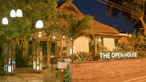 Un restaurante llamado "The Open House" con techo de paja, mesas al aire libre y un letrero que indica una siesta para tomar.